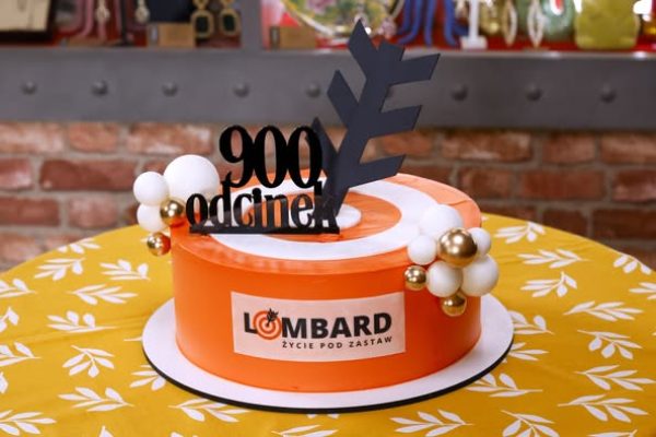 plan, plan serialu, Plan serialu "Lombard. Życie pod zastaw", Telewizja Puls, baloniki dekoracyjne, okrągły tort, dekoracja z napisem, napis Lombard Życie pod zastaw, pomarańczowy tort, ciasto okolicznościowe, biało-złote dekoracje, stół z żółtym obrusem, napis 900 odcinek, sierpień 2025, Czadoman w 900. odcinku "Lombard. Życie pod zastaw"